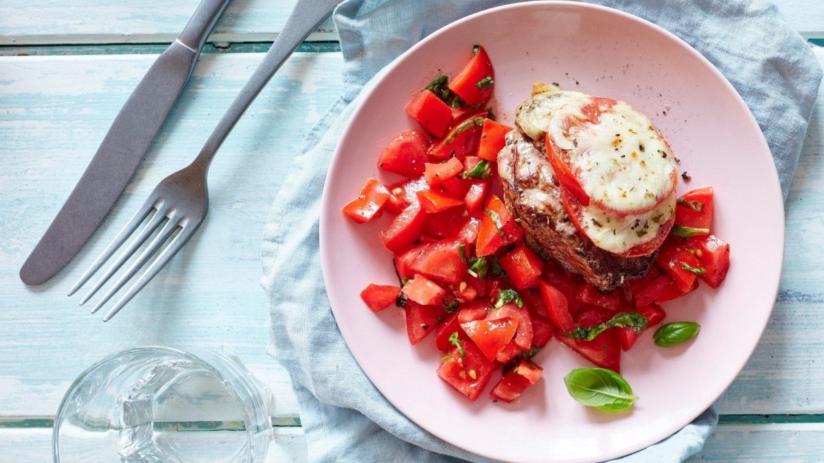 Unser Pizzaiola-Steak liegt fein angerichtet mit Tomatensalat auf einem rosa Teller zum Verzehr bereit.