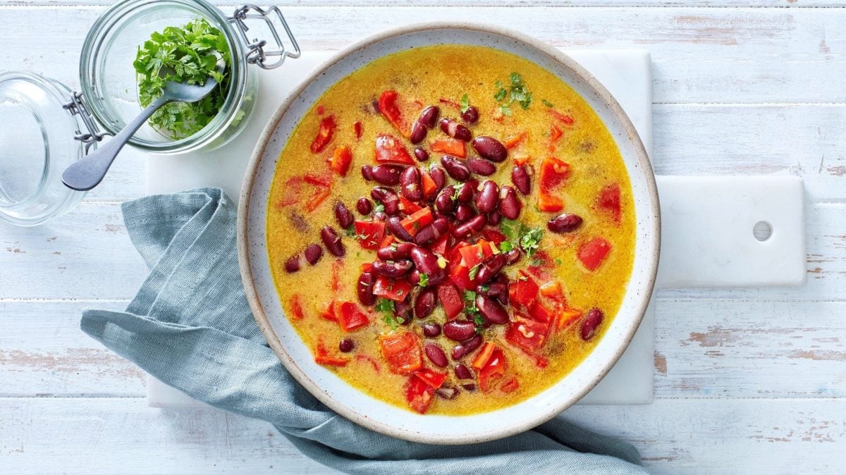 Ein Teller unserer scharfen Bohnen-Kokos-Suppe steht auf einem weißen Brett auf einem Holztisch. Petersilie steht als Garnitur daneben.