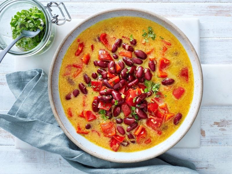 Ein Teller unserer scharfen Bohnen-Kokos-Suppe steht auf einem weißen Brett auf einem Holztisch. Petersilie steht als Garnitur daneben.