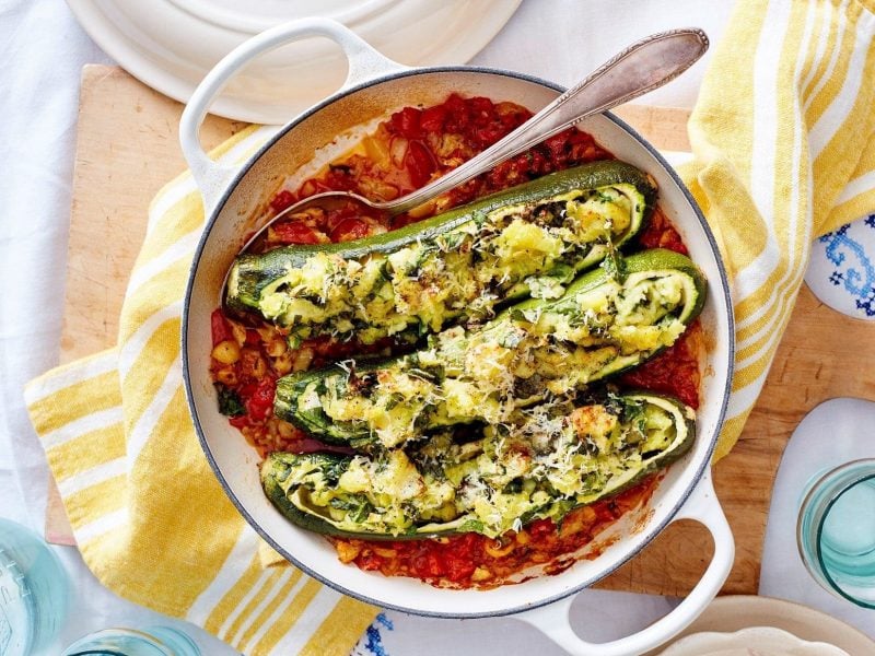 Drei Zucchini-Kartoffel-Schiffchen liegen in der Sauce in einem Bräter. Dieser steht auf einem Holzbrett auf dem Tisch.