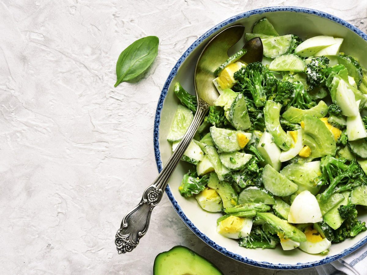 Saftiger Avocadosalat mit Brokkoli, Gurke und Ei mit einer feinen Dill-Sauce und Basilikumblättern in einer hellen Schale auf einem Steintisch serviert.