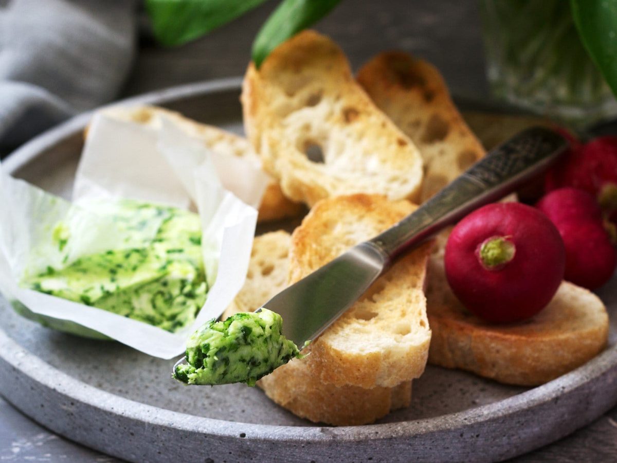 Bärlauch-Butter in Papier auf grauem Teller mit Baguette-Scheiben und Radieschen. Auf dem Brot ein Messer mit Bärlauch-Butter- Im Hintergrund graues Tuch. Frontalansicht.