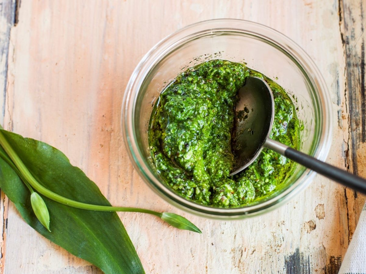 Bärlauch-Pesto in einem Glas mit silbernem Löffel. Daneben frischer Bärlauch. Angerichtet auch Holzbrett. Links am Bildrand blau-weißes Leinentuch. Aufnahme aus der Vogelperspektive.