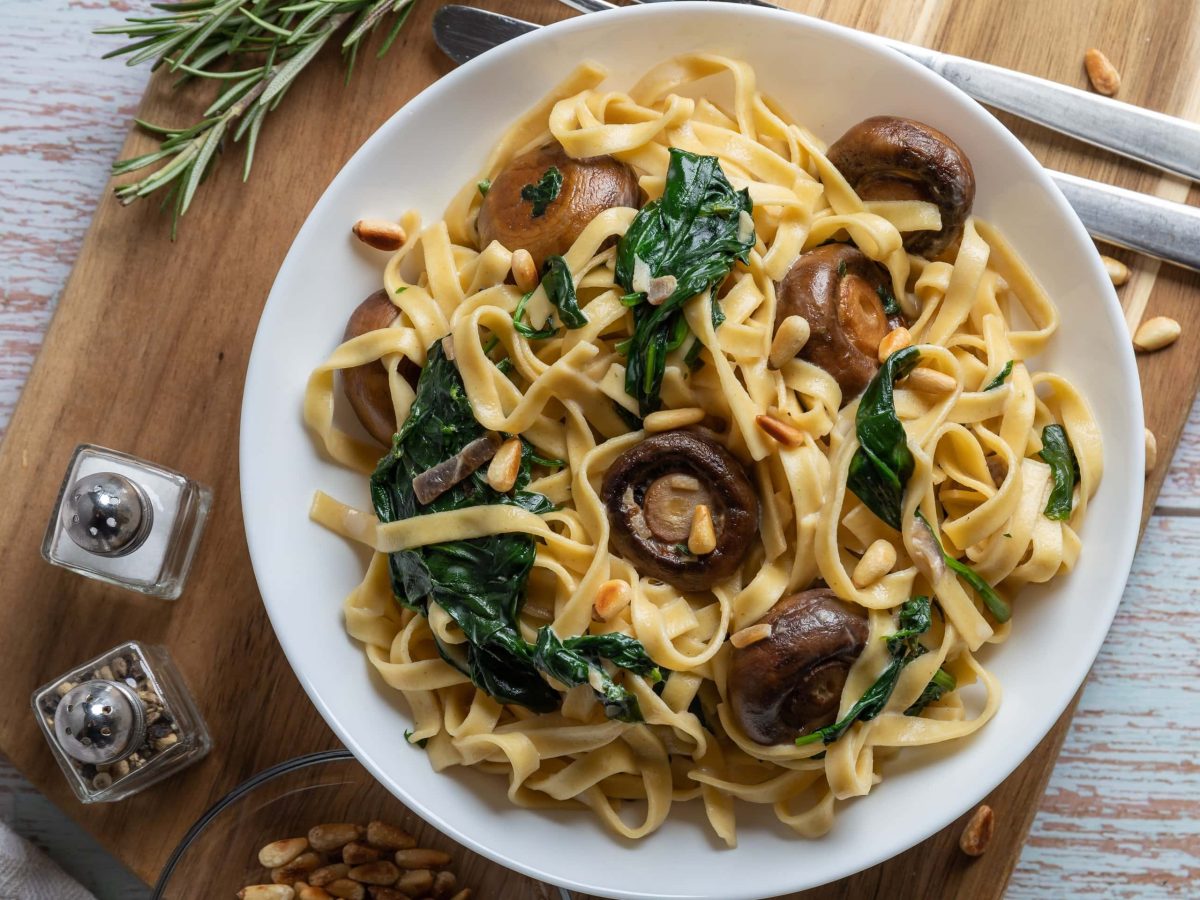 Ein Teller Bärlauch-Spaghetti mit Champignons und Pinienkernen auf einem Brett in der Draufsicht, daneben Gewürzstreuer und Besteck.