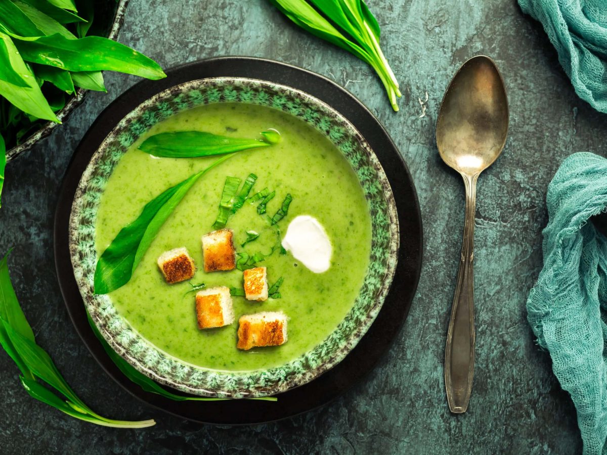 Ein Teller Bärlauchsuppe mit Croutons in der Draufsicht, daneben ein Löffel, Geschirrtuch und frischer Bärlauch.