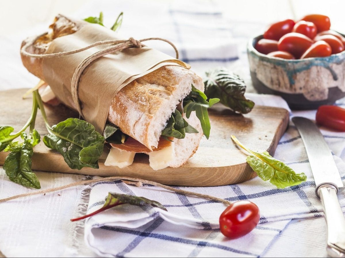 Auf einem weißen Untergrund aus Holz liegt ein Baguette mit Serrano-Schinken, Käse und Spinat. Neben diesem liegen ein Messer, Spinatblätter sowie mehrere frische kleine Tomaten.