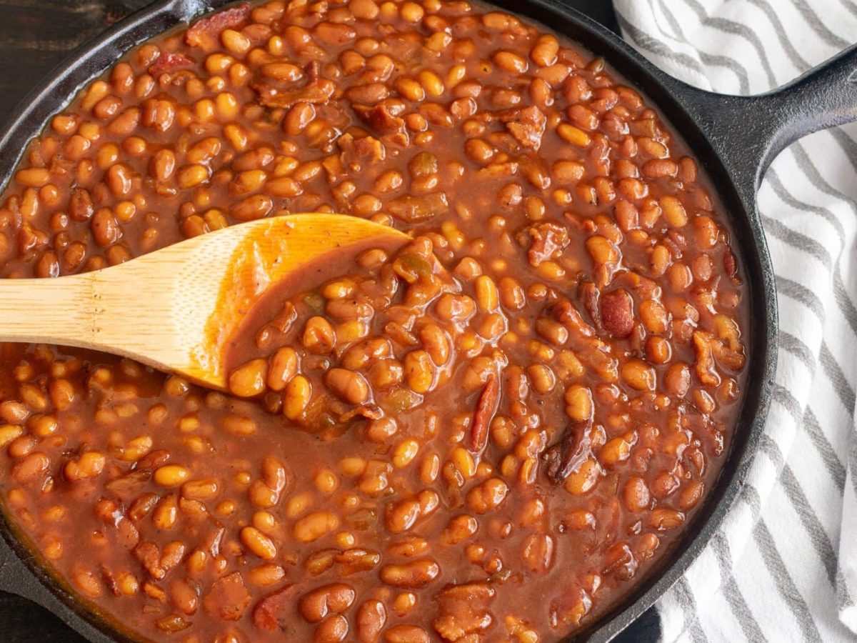 Selbst gemachte Baked Beans Heinz Beanz in schwarzer Guss-Pfanne auf dunklem Untergrund mit Holzlöffel. Daneben gestreiftes Stofftuch.