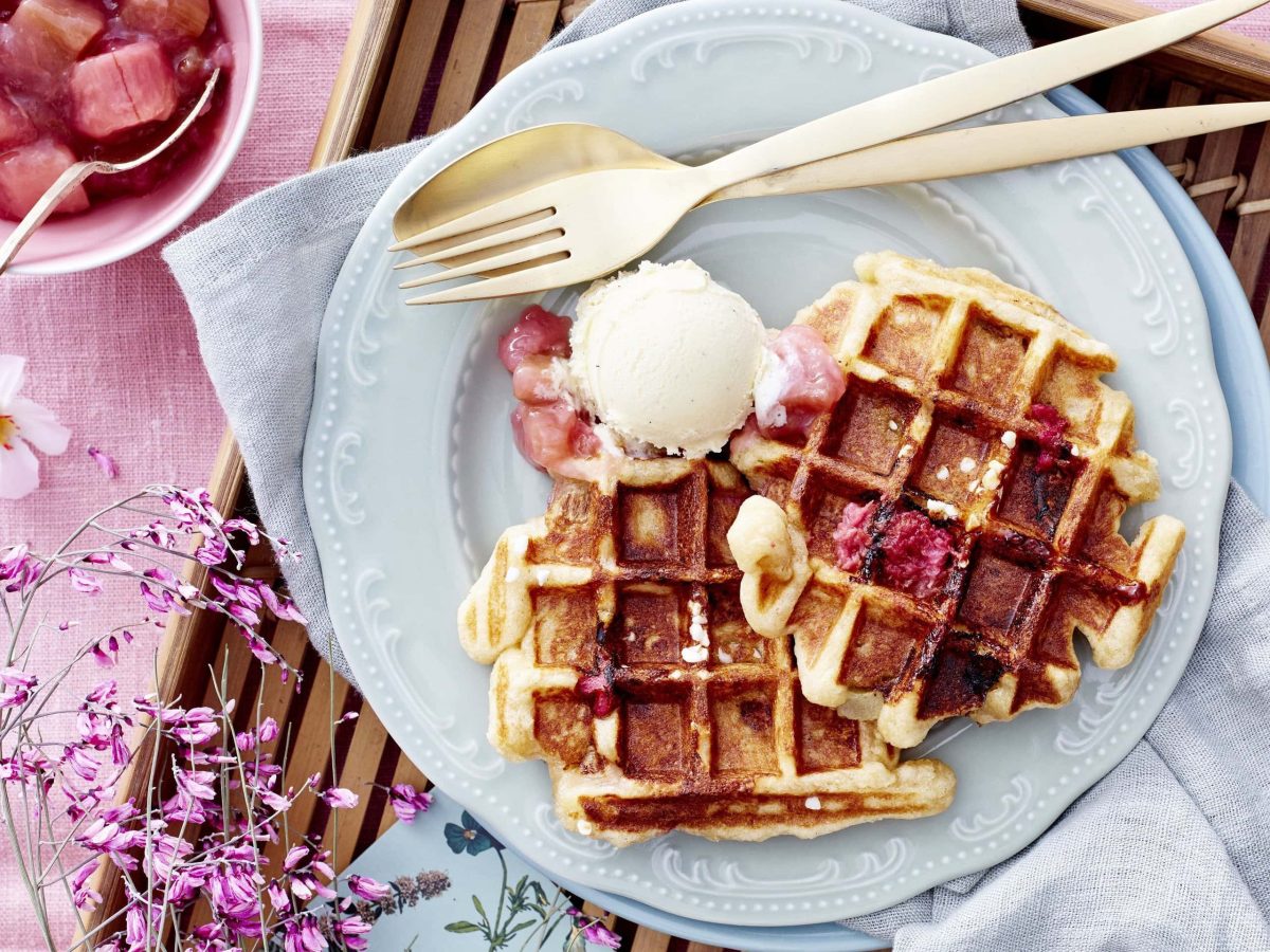 Belgische Rhabarber-Waffeln mit fruchtigem selbstgemachten Rhabarberkompott und einer Kugel Eis auf einem hellen Teller auf einem mit rosaner Tischdecke verzierten Tisch.