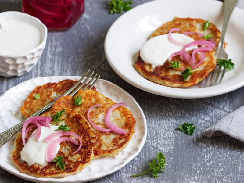 Saftige Boxty-Pancakes, bestehend aus Pfannkuchenteig mit Sahne und geriebener Kartoffel, serviert mit Schmand, eingelegten Zwiebeln in Essig und frischer Petersilie auf hellen Tellern auf hölzernem Untergrund.