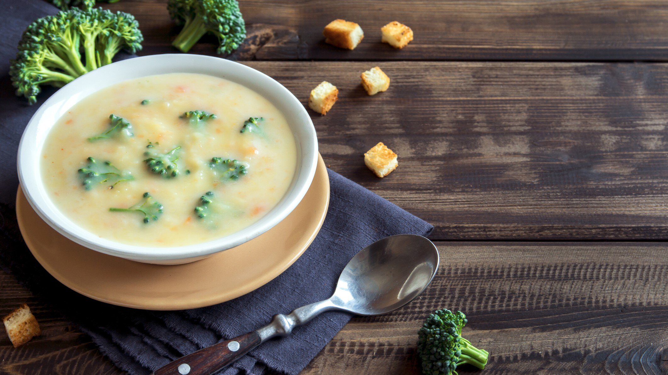 Brokkoli Cheddar Suppe auf einem weißen Teller. Daneben verschiedene Zutaten, Küchentücher und ein Löffel. Das Ganze auf einem hölzernen Untergrund.