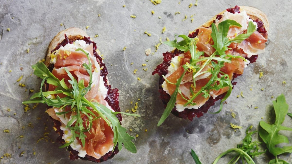 Zwei Scheiben Bruschetta mit roter Bete, daneben etwas Rucola. Das Ganze auf einem grauen Untergrund.