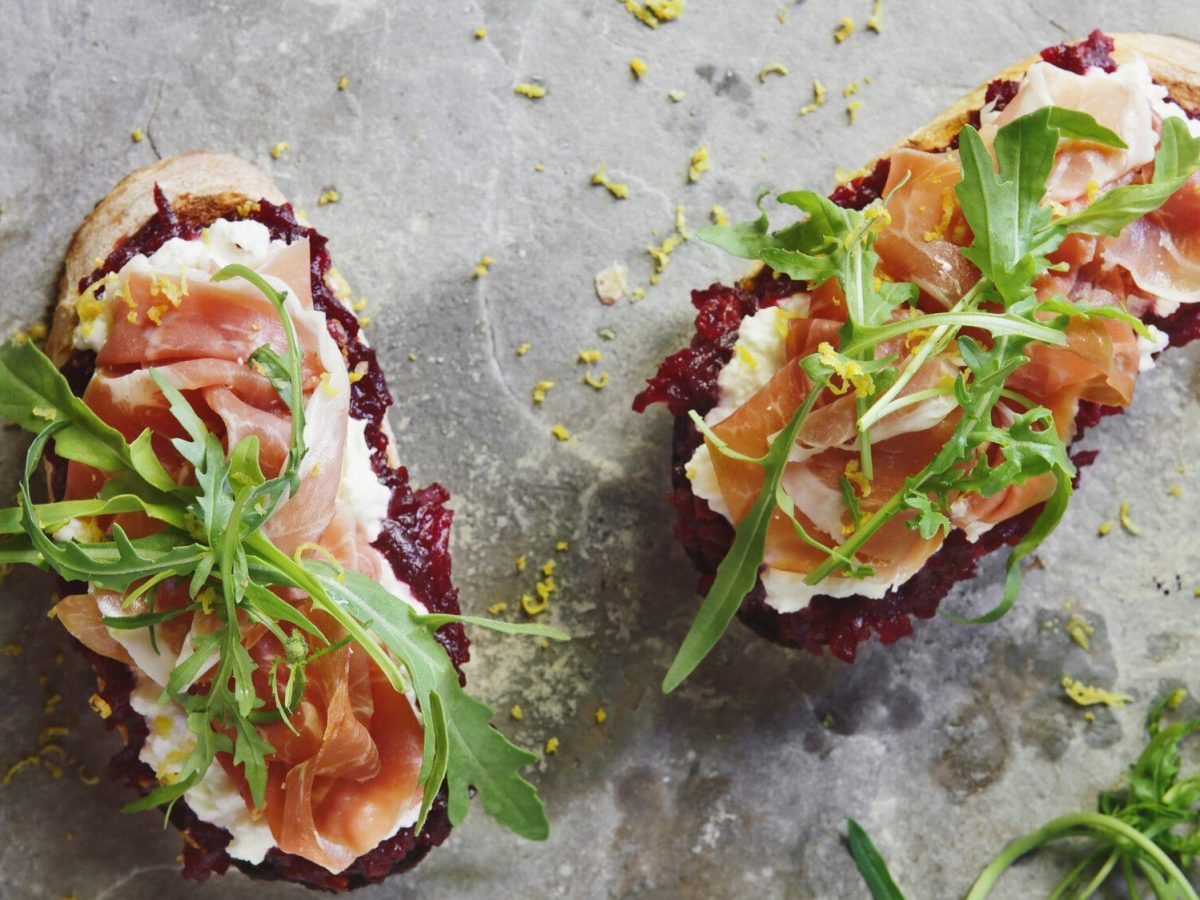 Zwei Scheiben Bruschetta mit roter Bete, daneben etwas Rucola. Das Ganze auf einem grauen Untergrund.