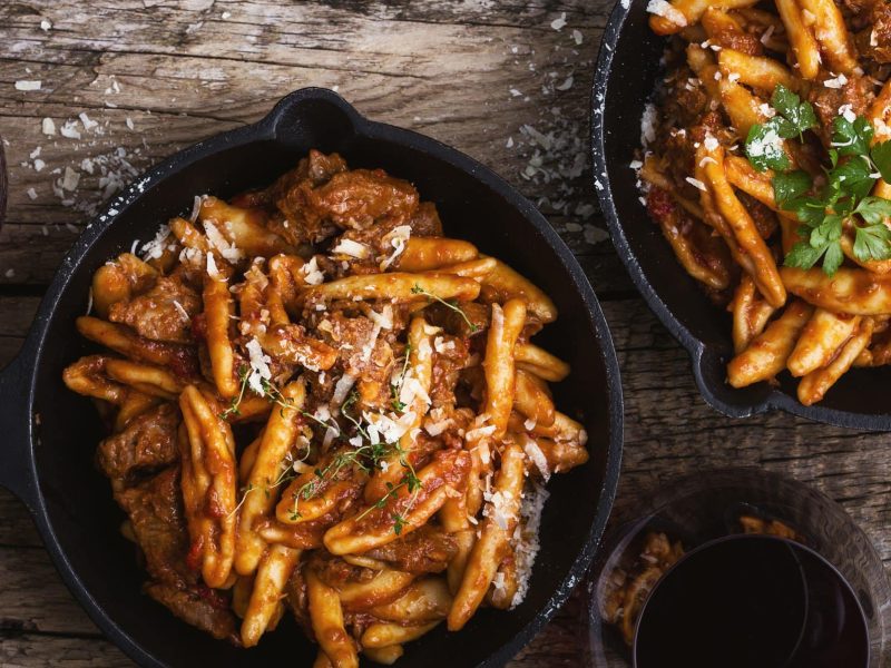 Casareccia Pasta mit Rinderragout in zwei gusseisernen Pfannen. Das Ganze auf einem hölzernen Untergrund.