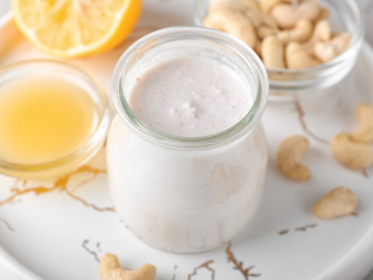 Ein Glas Cashewsahne auf einem weißen Tablett, daneben eine Schale Cashewkerne und eine Schale Zitronensaft.