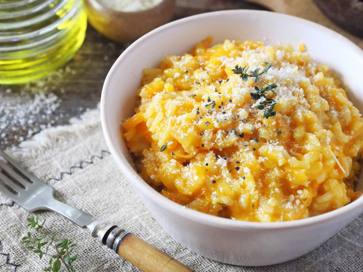 Cheddar Risotto in einer weißen Schüssel. Daneben einige Zutaten sowie eine Gabel. Das Ganze auf einem Küchentuch auf einem hölzernen Untergrund.