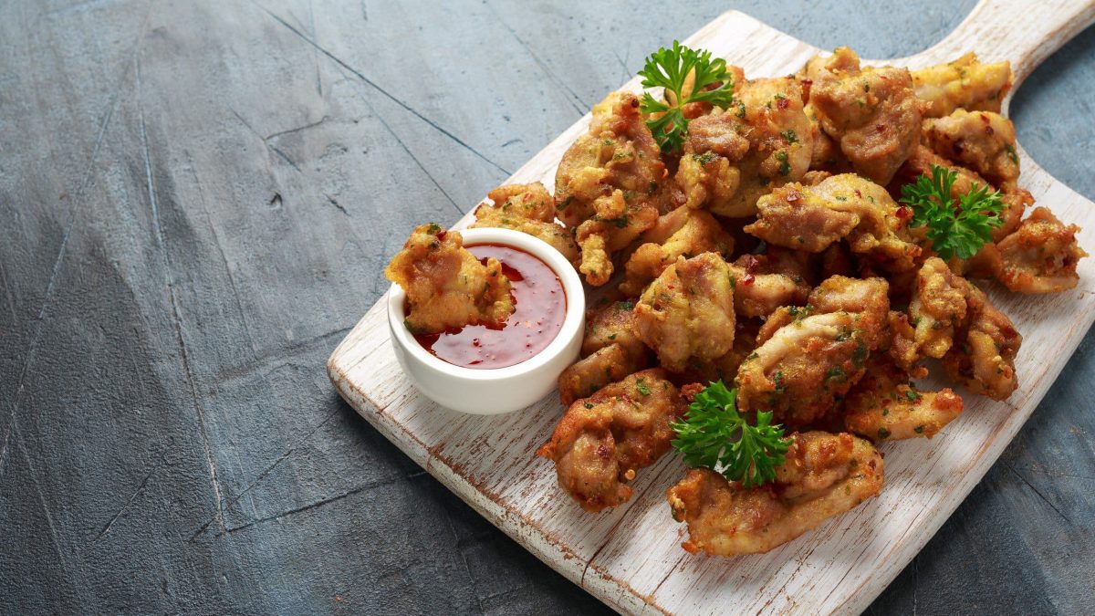 Chicken Pakora indisches Fried Chicken auf Holzplatte mit rotem Dip auf grauem Untergrund. Koriander als Dekoration verteilt. Aufnahme aus der Vogelperspektive.