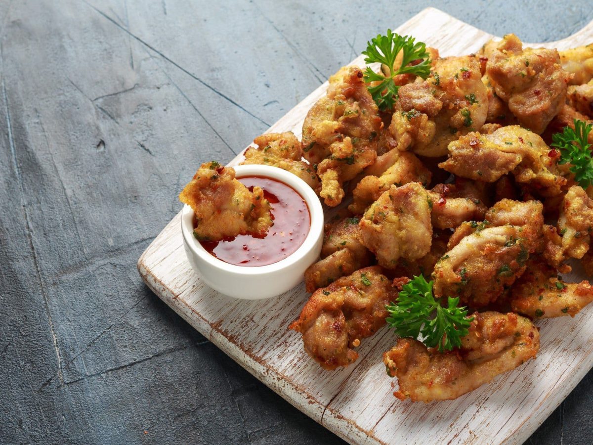 Chicken Pakora indisches Fried Chicken auf Holzplatte mit rotem Dip auf grauem Untergrund. Koriander als Dekoration verteilt. Aufnahme aus der Vogelperspektive.