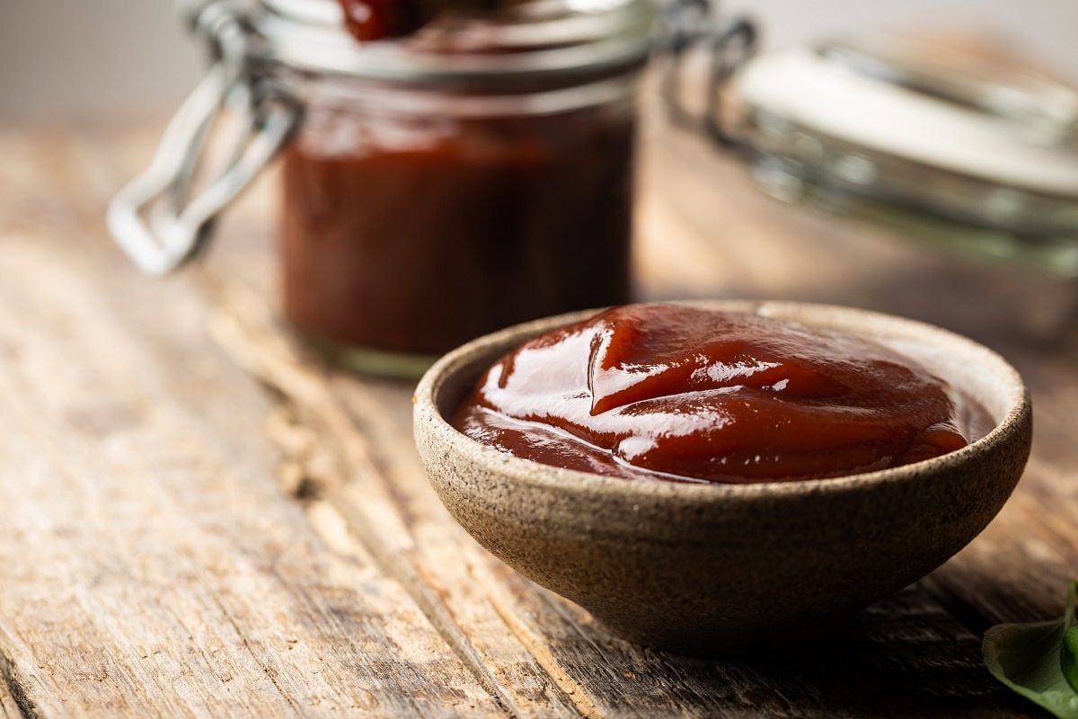 Dr. Pepper Barbecue-Sauce in brauner Schale auf Holztisch. Im Hintergrund Sauce im Einweckglas. Frontalansicht.