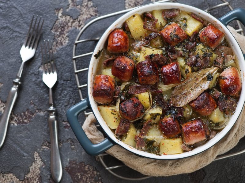 Saftiger Dublin Coddle mit Würstchen, Bacon und Kartoffeln in einer würzigen Biersauce, serviert in einem großen Topf auf steinernem Untergrund.