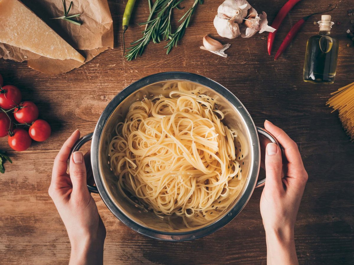 Ein Topf Spaghetti in den Händen einer Frau in der Draufsicht, drumherum liegen auf einem Holztisch Gemüse, Parmesan, Nudeln und andere Zutaten.