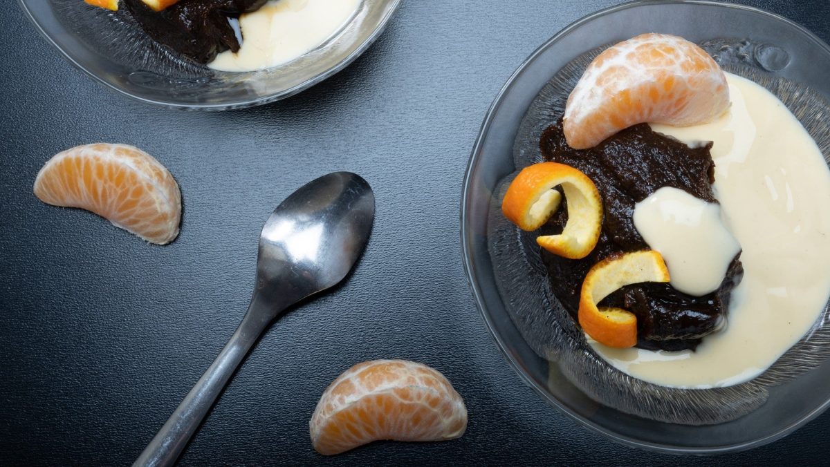 Finnischer Mämmi Malz-Pudding in Glasschale mit Vanillesauce, Mandarinen-Spalten und Mandarinenschale auf grauem Tisch. Daneben Teelöffel, Mandarinen-Spalten und eine weitere Glasschale. Aufnahme aus der Vogelperspektive.