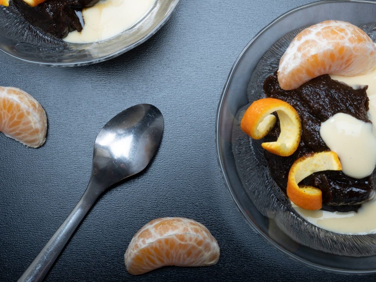 Finnischer Mämmi Malz-Pudding in Glasschale mit Vanillesauce, Mandarinen-Spalten und Mandarinenschale auf grauem Tisch. Daneben Teelöffel, Mandarinen-Spalten und eine weitere Glasschale. Aufnahme aus der Vogelperspektive.