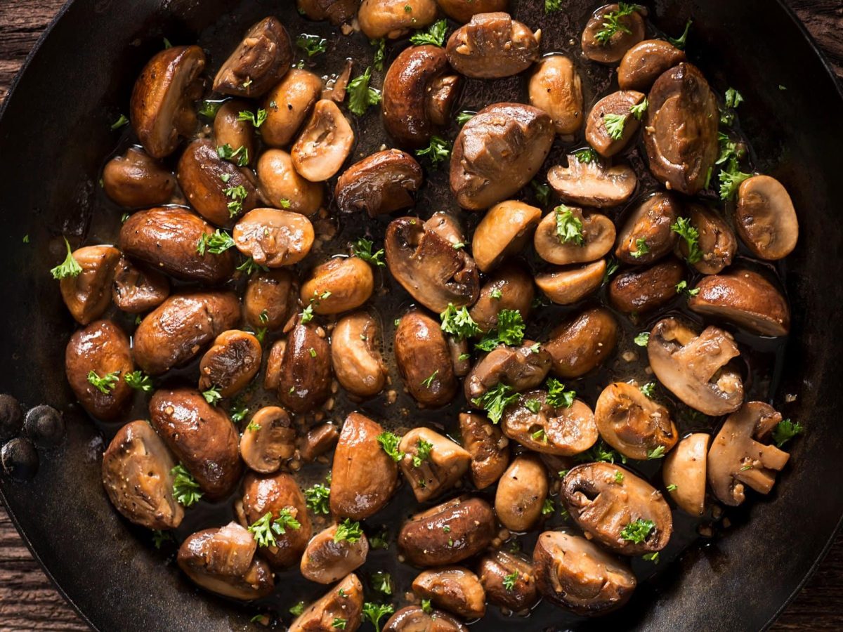 Gebratene Champignons mit Zwiebeln in gusseiserner Pfanne auf Holzfläche.