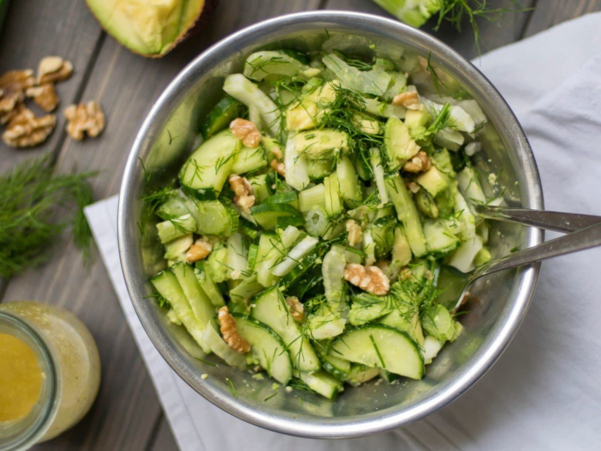 Eine Schüssel Gurkensalat mit Fenchel und Walnüssen mit Salatbesteck in der Draufsicht, daneben Zutaten wie Nüsse, Fenchelgrün, Gurke und Dressing.