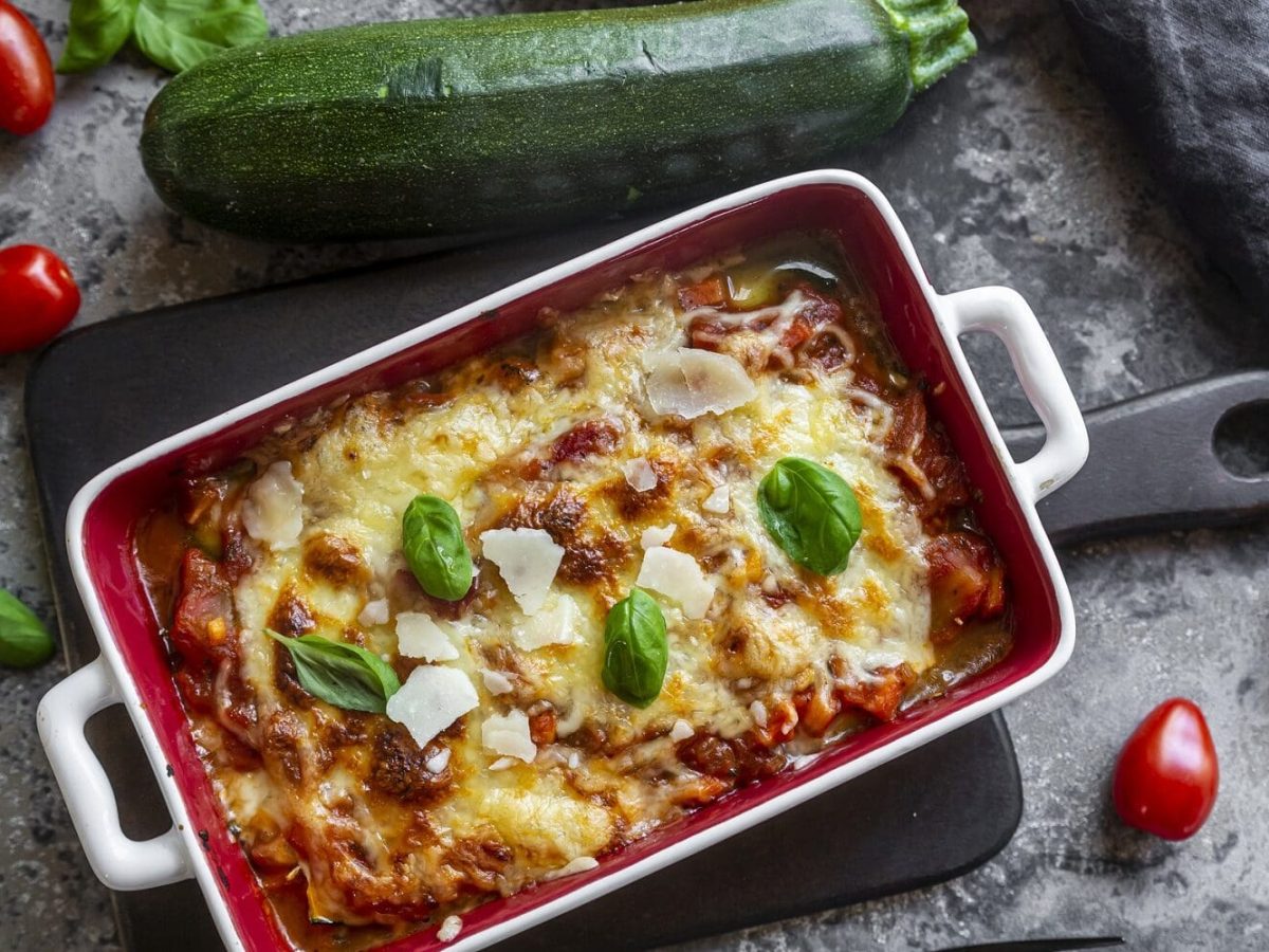 Aus der Vogelperspektive: Auf einem grauen Untergrund liegen eine Zucchini, TOmaten und Bassilikum-Blätter. Und es steht eine Hackfleisch-Zucchini-Lasagne in einer großen Form auf dem Untergrund.