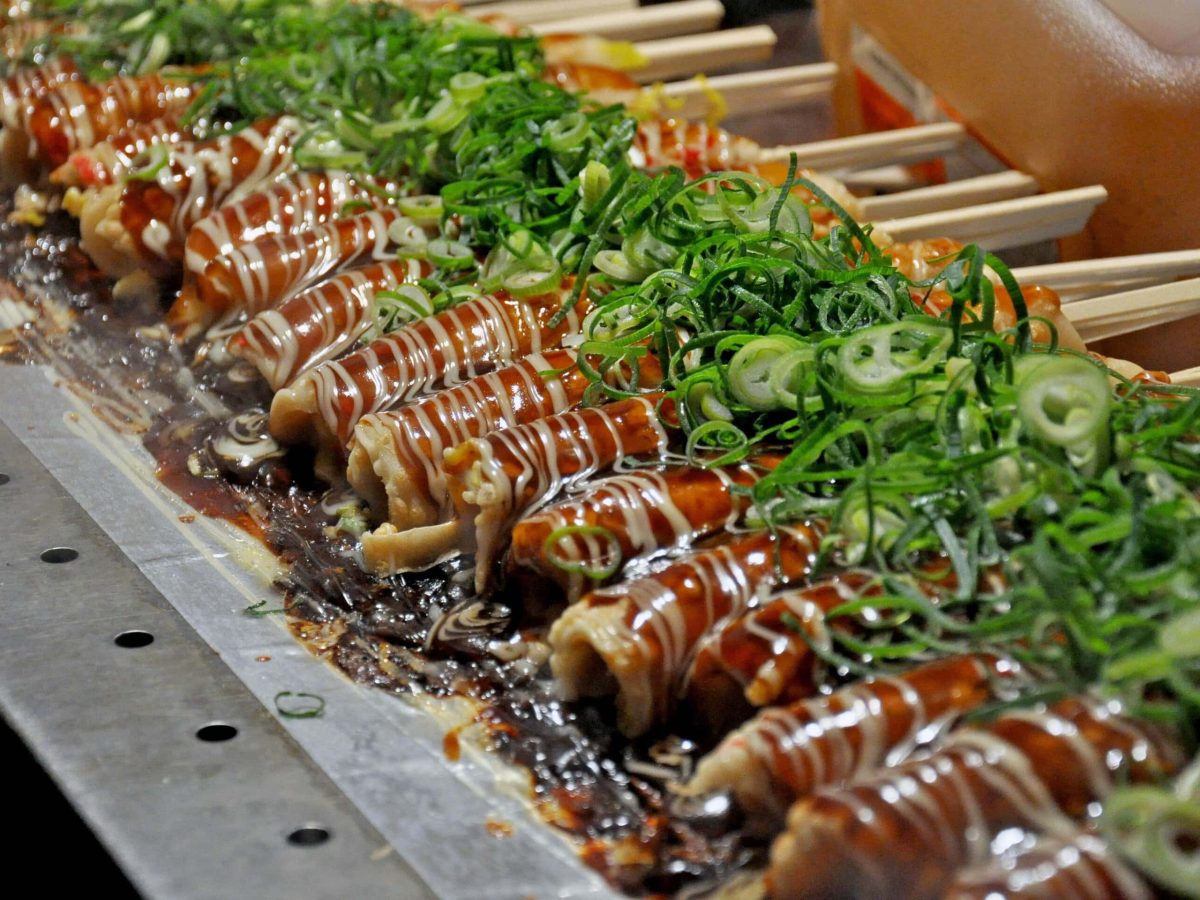 Hashimaki auf Gasgrill aus Metall an Streetfood-Stand in Japan. Im Hintergrund Kanister mit Öl. Frontalansicht.