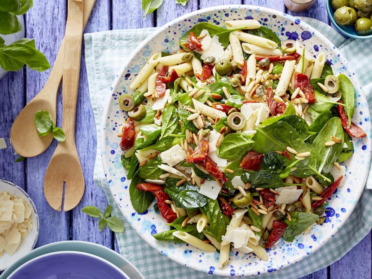 Italienischer Nudelsalat mit Oliven und frischem Basilikum mit Parmesan auf einem Teller auf einem gedeckten Tisch neben zwei Kochlöffeln