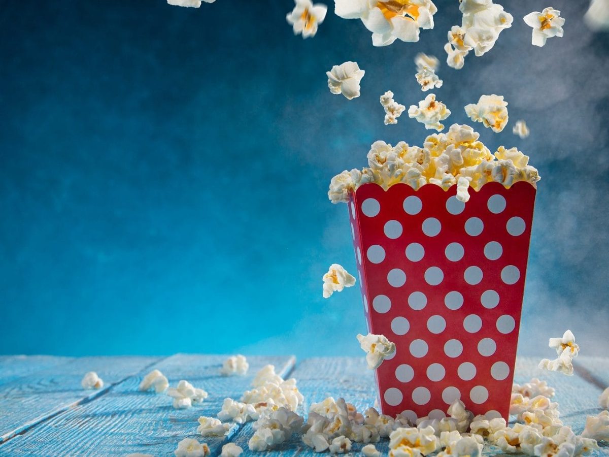 vor einem blauen hintergrund ist eine rote weiße gepunktete popcorntuete zu sehen. aus der springen popcornkoerner. das foto bebildert das buch Kochen in Perfektion : Profi-Wissen für die Kueche