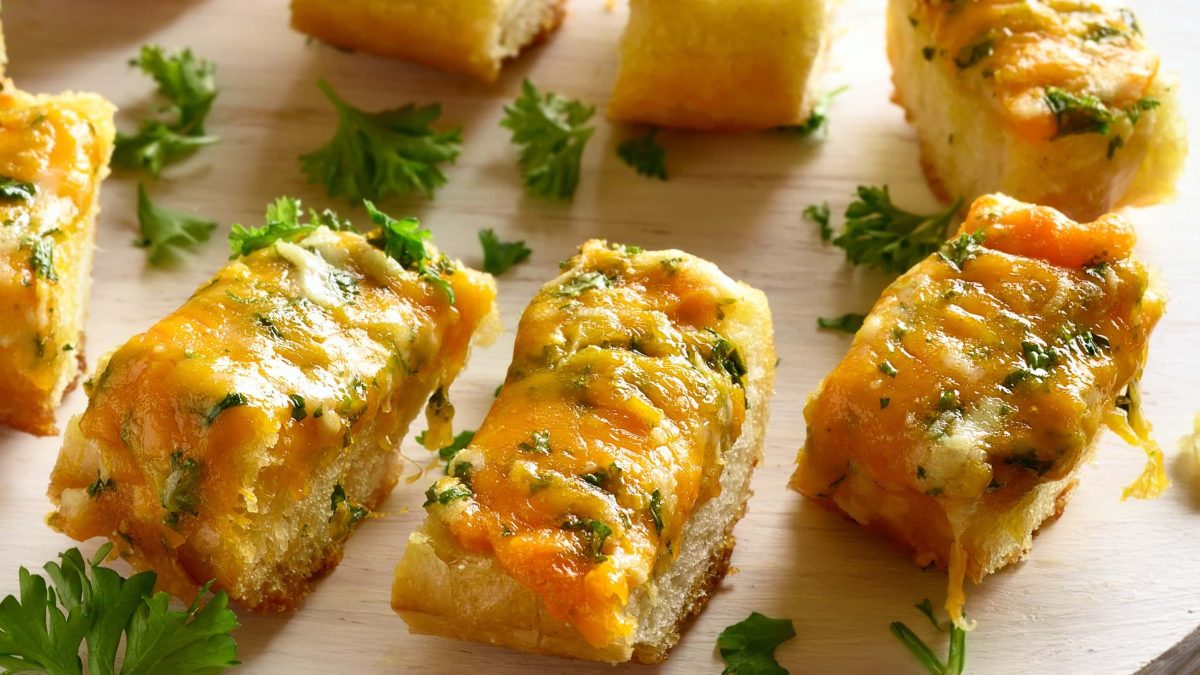 Mehrere Stücken Kräuterbaguette mit Käse liegen auf einem hellen Holzbrett. Die Baguettestücken sind angerichtet mit Petersilie.