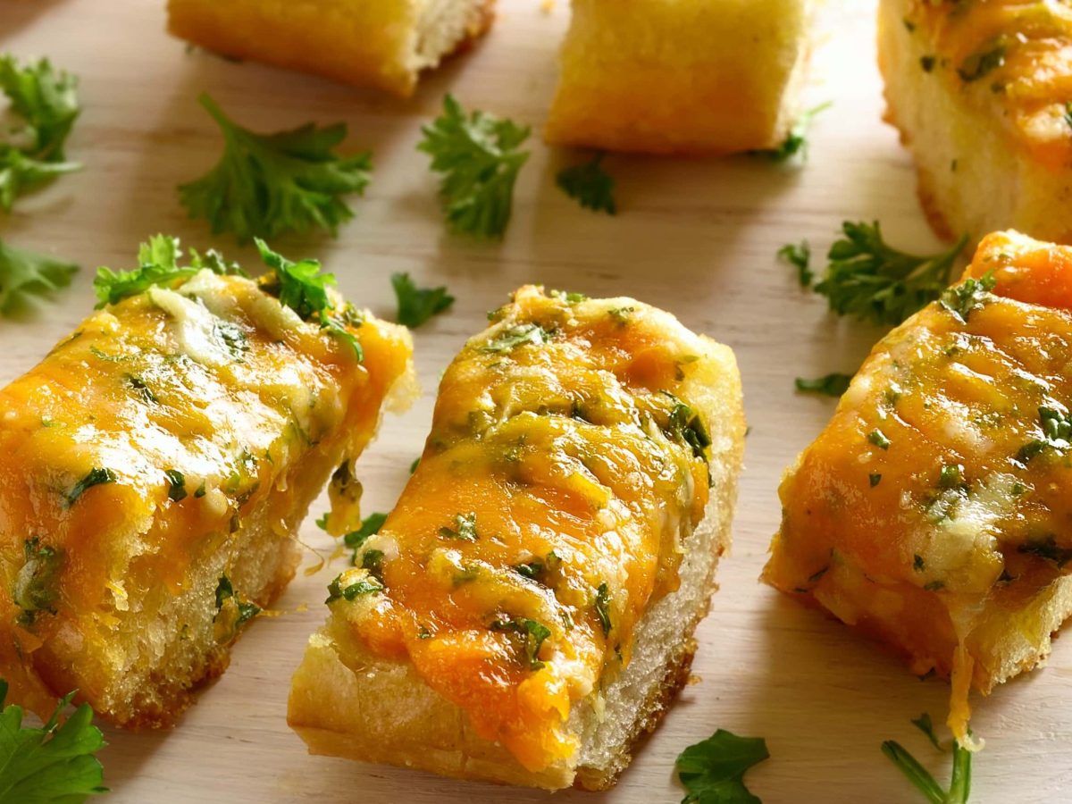 Mehrere Stücken Kräuterbaguette mit Käse liegen auf einem hellen Holzbrett. Die Baguettestücken sind angerichtet mit Petersilie.