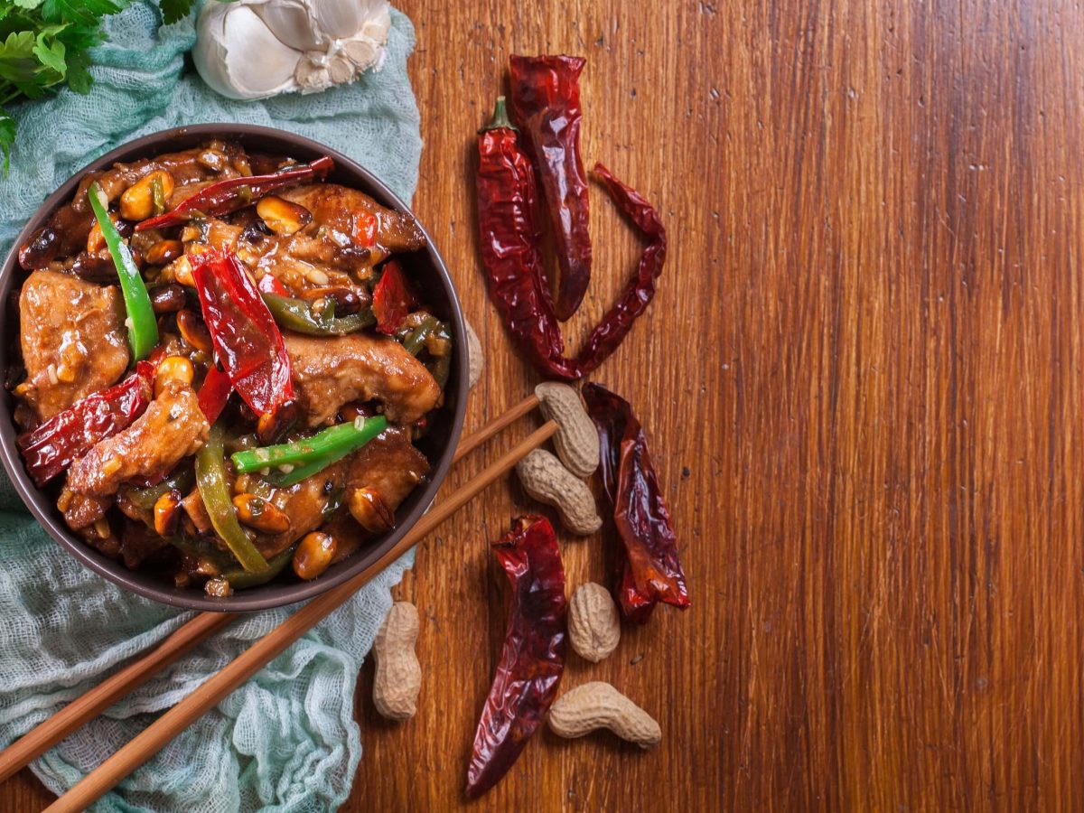 Kung Pao Chicken in schwarzer Schüssel auf blauem Tuch auf Holztisch. Daneben getrocknete Chilis, Essstäbchen, Knoblauch und Koriander. Aufnahme aus der Vogelperspektive.