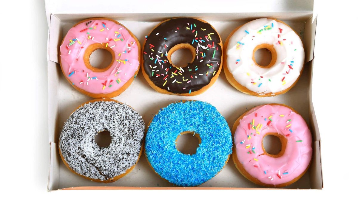 Sechs verschiedene Donuts liegen in einer weißen Box auf einem weißen Untergrund. Zwei Donuts sind mit rosa Glasur überzogen, einer mit blauen Streuseln, einer mit Schokolade, ein weiterer Donut mit Schokolade und Kokosraspeln und ein letzter mit weißem Zuckerguss.