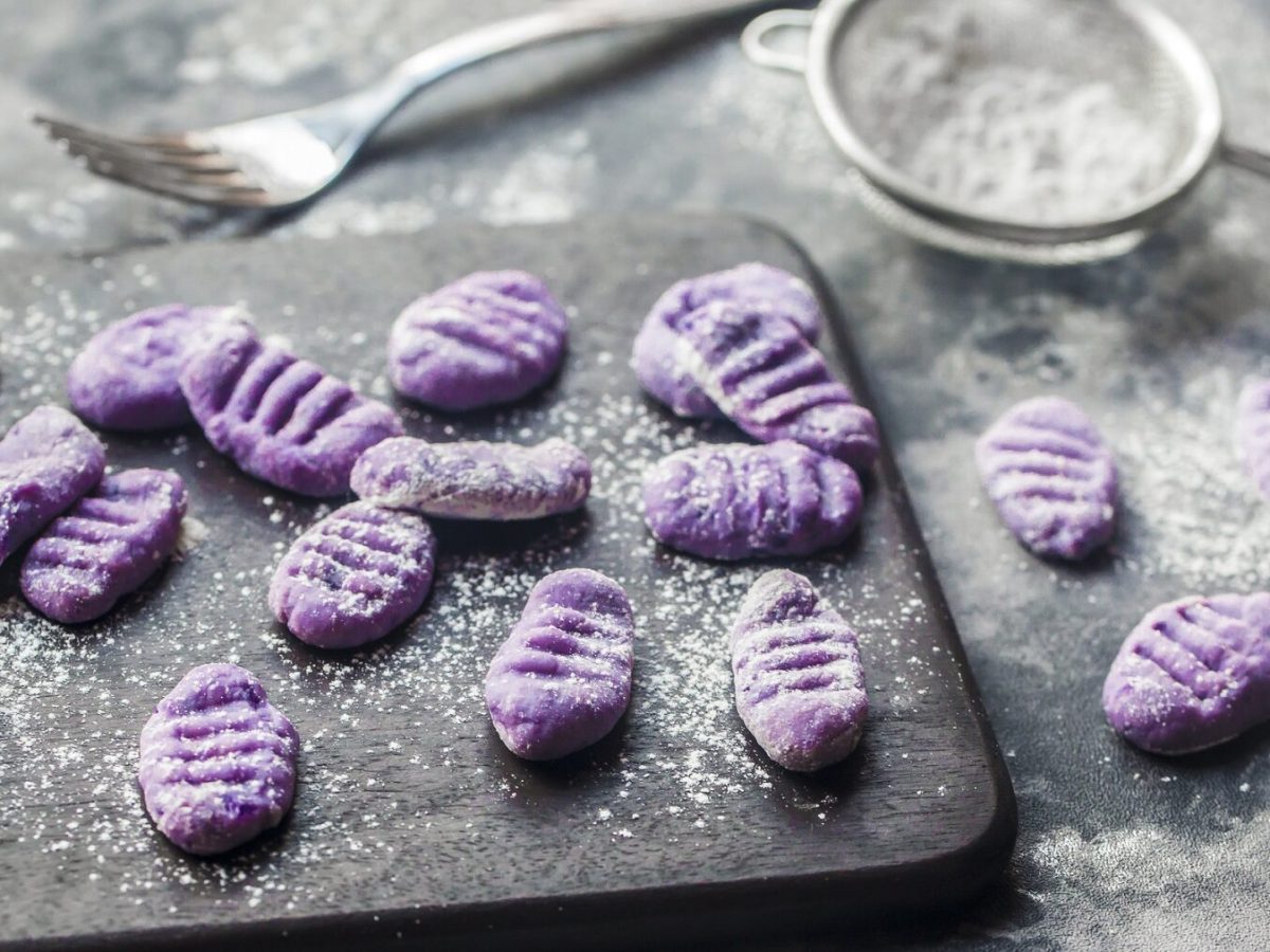 Aus der Vogelperspektive: Auf einer schwarzen Schieferplatte liegen Lila-Kartoffel-Gnocchi, eine Gabel und ein kleines Sieb mit einem weißen Pulver. Es handelt sich um Mehl-