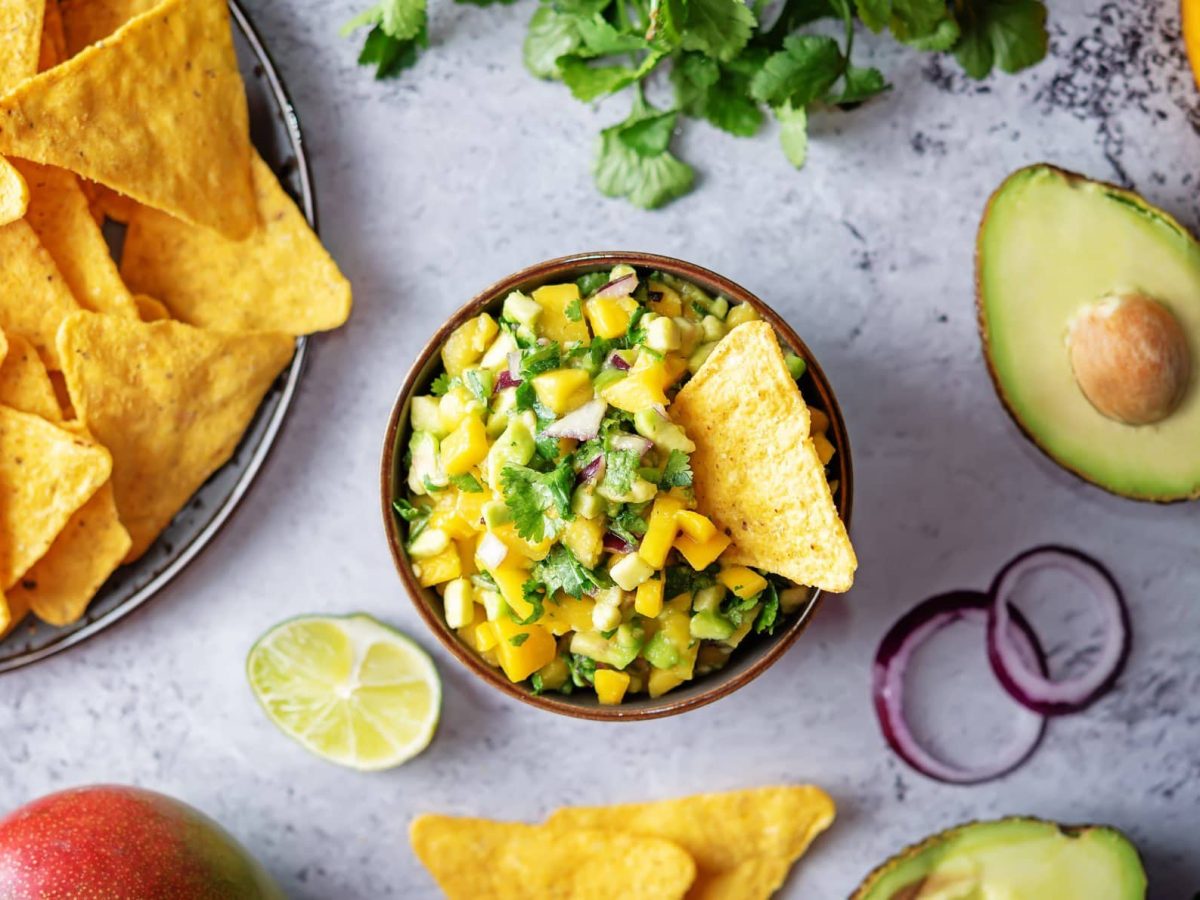 Eine Schüssel Mango-Avocado-Salsa auf grauem Beton in der Draufsicht, daneben liegen Nachos und Zutaten wir Avocado, Mango und Kräutern.