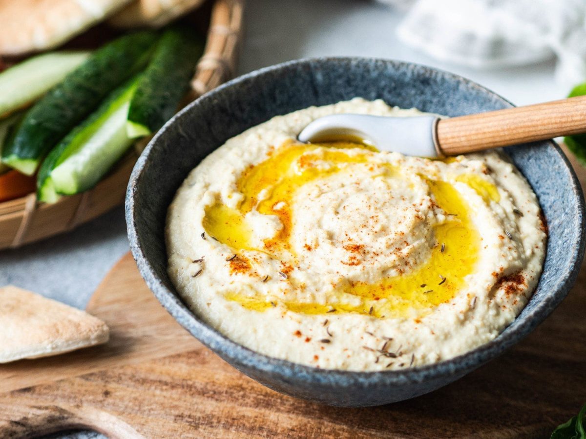 Eine Schale Mangold-Hummus mit Öl und einem Holzlöffe, im Hintergrund Germüse.