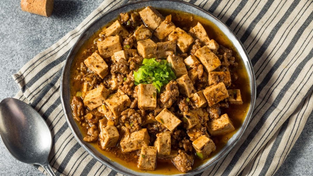 Ein Teller Mapo-Tofu mit klein geschnittener Frühlingszwiebeln auf einem Geschirrtuch in der Draufsicht, daneben liegt ein Löffel.