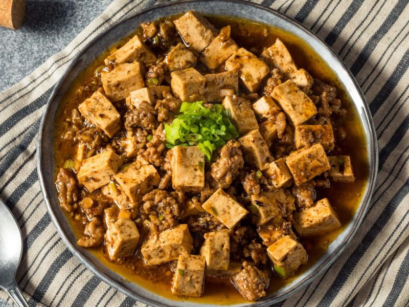 Ein Teller Mapo-Tofu mit klein geschnittener Frühlingszwiebeln auf einem Geschirrtuch in der Draufsicht, daneben liegt ein Löffel.