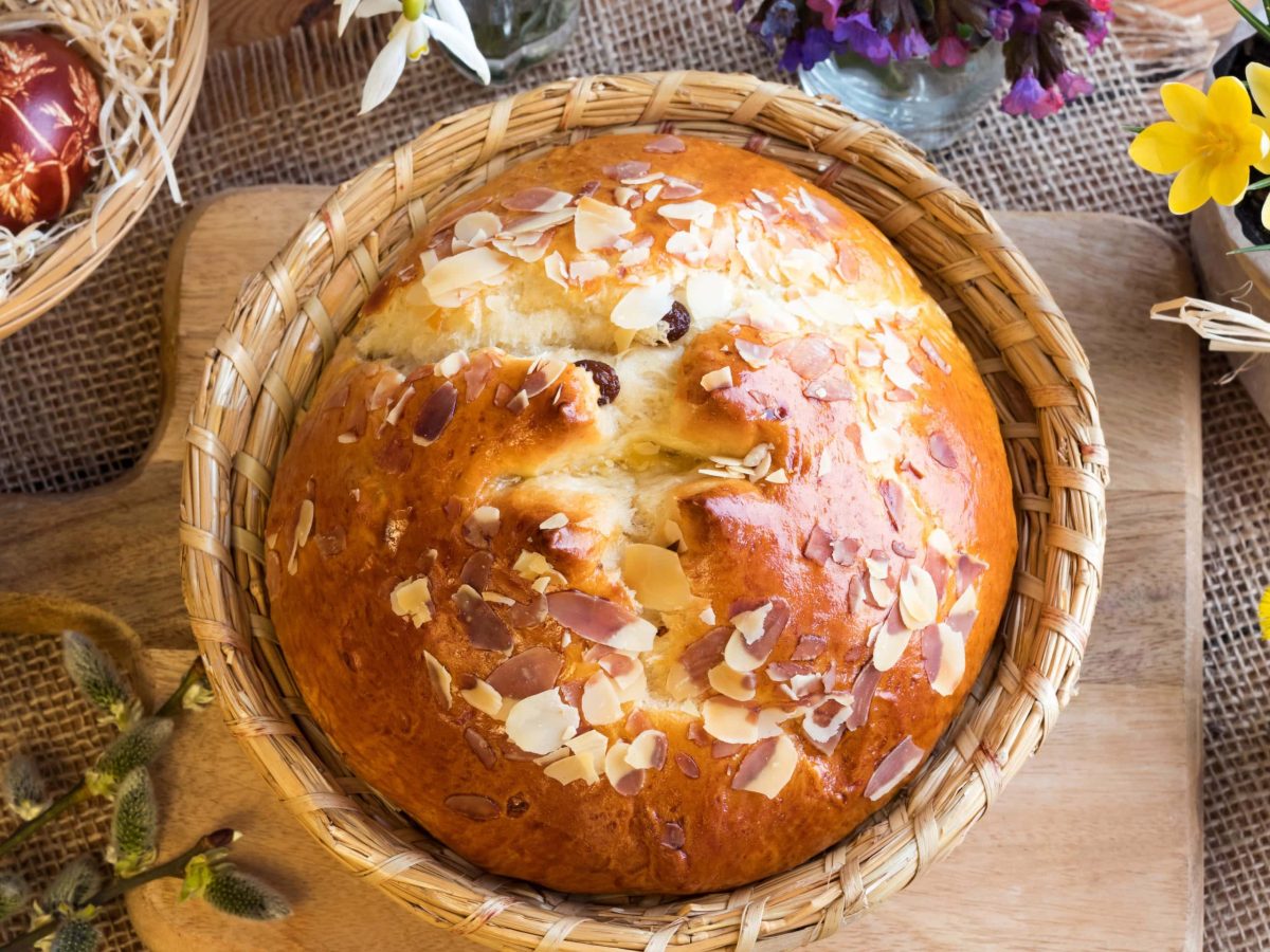 Böhmisches Mazanec Osterbrot mit Rum-Rosinen in Osterkorb auf Holzbrett und Holztisch. Gelbe Blumen und bemalte Eier in Osternest als Deko im Bild. Aufnahme aus der Vogelperspektive.