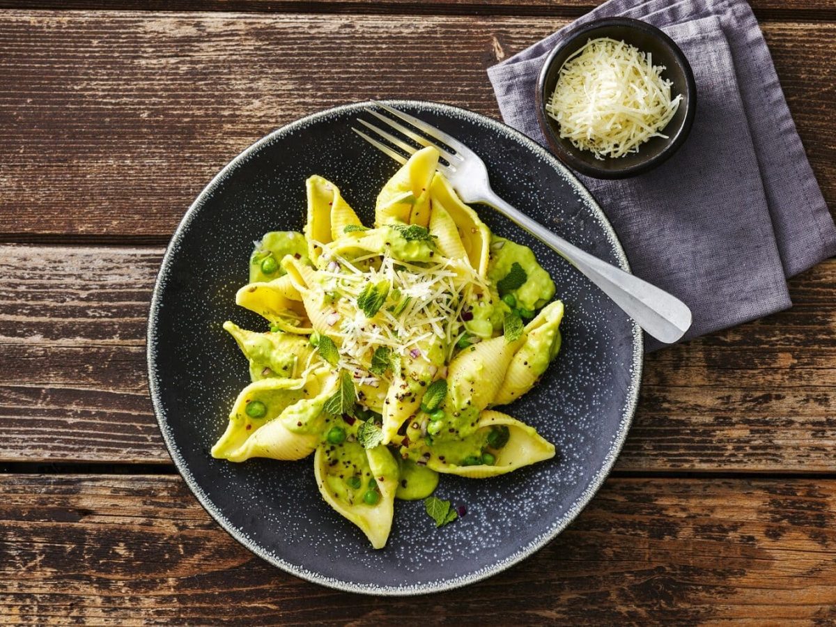 Aus der Vogelperspektive: Auf Holzdielen ist ein großer dunkler Teller zu sehen. In dem sind Muschelnudeln mit Avocado-Erbsen-Minze-Füllung.