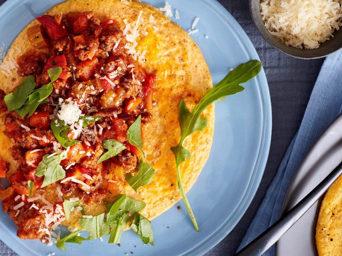 Ein Omelette mit Bolognese, Rucola und Parmesan auf einem blauen Teller in der Draufsicht. Daneben Besteck und eine Schüssel Parmesan.