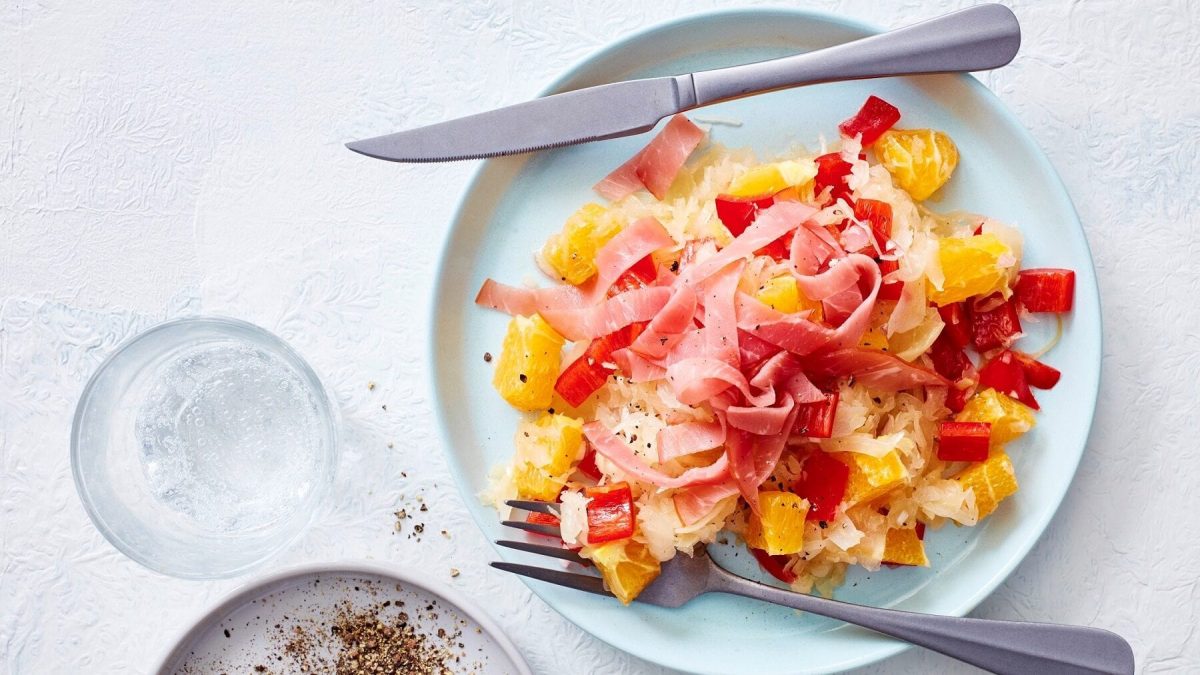 auf einem hellen Untergrund steht ein himmelblauer Teller. Auf dem Teller ist ein Orangen-Sauerkraut-Salat. Neben dem Salat lieben ein Messer und eine Gabel. Vor dem Teller steht ein Glas Wasser