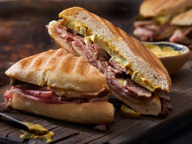 Zwei grilled Cuban Sandwiches auf dunklem Holzbrett auf Holztisch. Messer mit schwarzem Griff und Senf im Vordergrund. Rechts unscharf weiteres Sandwich. Frontalansicht.