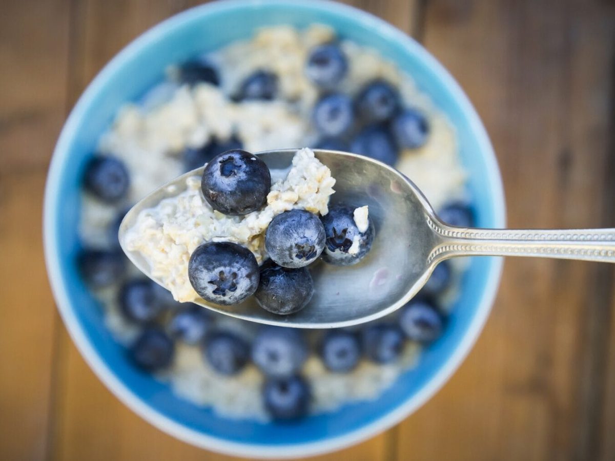 Aus der Vogelperspektive: Ein Löffel mit Overnight Oats mit Blaubeeren. wird über eine Schüssel mit der Frühstücksmahlzeit gehalten.