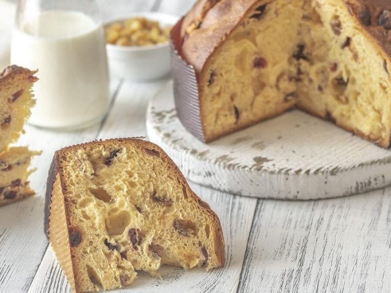 Auf einem weißen Untergrund aus Holz liegen zwei Scheiben Panettone. Dies ist ein italienischer Feiertagskuchen. Der restliche Panettone steht auf einer weißen Platte. Im Hintergrund ist eine Glasflasche mit einer weißen Flüssigkeit zu sehen. es handelt sich wahrscheinlich um Milch.