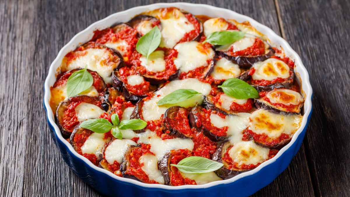 Ein Auflauf aus frittierter Aubergine mit überbackenen Käse, im italienischen genannt Parmigiana die Melanzane, in einer blauen Auflaufform auf einem hölzernen Tisch.