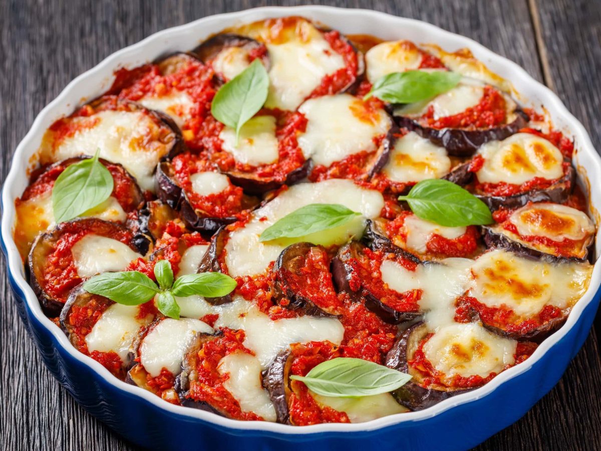 Ein Auflauf aus frittierter Aubergine mit überbackenen Käse, im italienischen genannt Parmigiana die Melanzane, in einer blauen Auflaufform auf einem hölzernen Tisch.